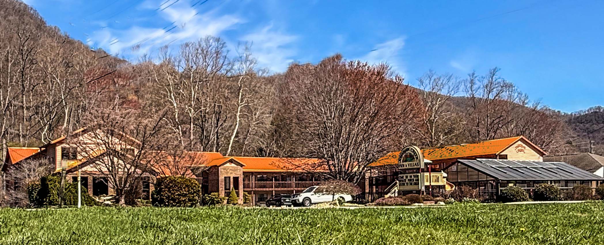 A long, rustic building with an orange roof sits in front of leafless trees and hills. A white car is parked near a sign, a glass greenhouse structure, and what could be a cozy Maggie Valley hotel. The grass in the foreground is green under a blue sky.